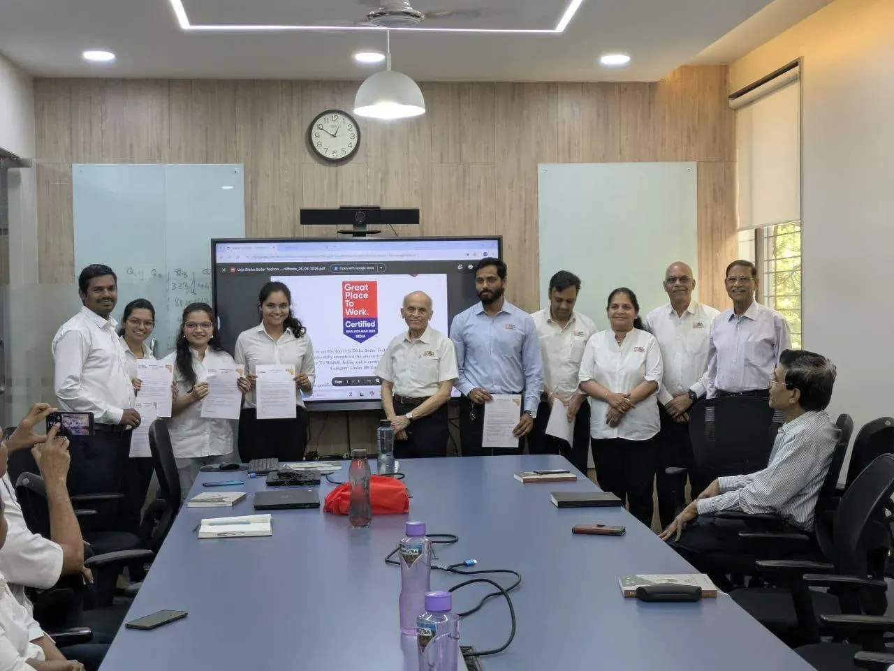 Urja-Disha employees holding their appreciation letters in the conference room, with the Great Place To Work certificate on the screen behind them