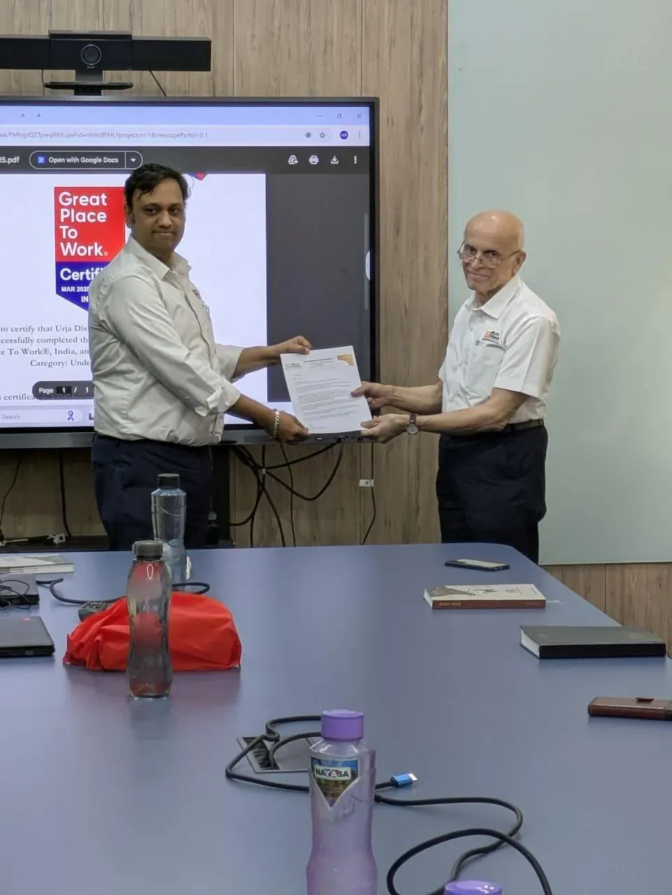 An employee being handed his appreciation letter in front of the Great Place To Work certificate shown on the conference room screen